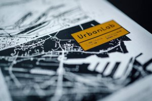 Ż&oacute;łty logotyp UrbanLab na tle czarno-białej grafiki miasta
