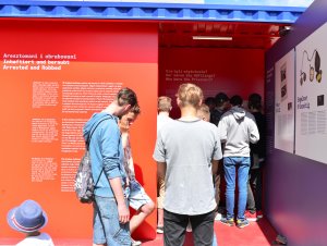Fragment kontenera w kolorach czerwonym i niebieskim z planszami wystawy z napisami i zdjęciami, w środku tłum młodych ludzi