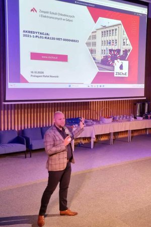 Mężczyzna przemawiajacy przed widownią z mikrofonem w ręku, wdalszym planie prezentacja