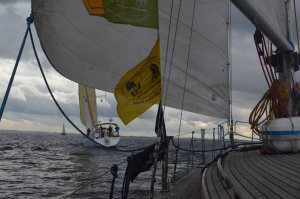 Zdjęcie z parady jachtów, która odbyła się w ramach zlotu "Próchno i Rdza". Na pierwszym planie fragment drewnianego pokładu jednego z jachtów i żagle powiewające na wietrze. W tle inny płynący jacht // fot. E. Banaszek