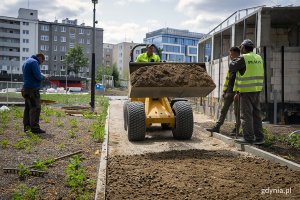 Koparka wiezie piasek, obok stoją robotnicy.