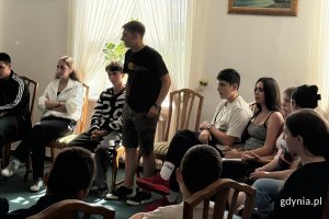 Przedstawiciel młodzieży z Gruzji w krótkich spodniach i czarnej koszulce przemawia podczas spotkania sali wizytowej, na fotelach wokół niego siedzi młodzież.