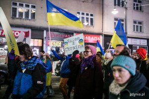 Mieszkańcy maszerują wieczorem ulicą podczas marszu antywojennego, trzymając flagi Ukrainy i hasła antywojenne.