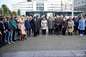 Uczestnicy uroczystości upamiętniających 86. rocznicę pierwszych wysiedleń gdynian zebrani przed tablicą pamiątkową na fasadzie budynku Dworca PKP Gdynia Główna