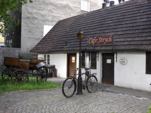 Zdjęcie z zewnątrz. Kamienny budynek z drewnianymi małymi okienkami.Na przeciwko wejścia znajduje się stary rower. Nad wejściem szyld "Cafe Strych".