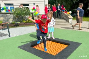 Chłopiec na placu zabaw, na trampolinie, w tle na schodach przed budynkiem ludzie świętujący uroczystość