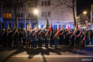 Grupa osób w biało-czerwonych szarfach i ze sztandarami stojąca przed pomnikiem