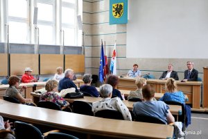 Seniorzy siedzą w ławach radnych miejskich na przeciwko trzech osób siedzących za pulpitem.