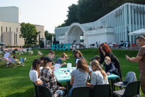 Grupa młodych ludzi siedzi przy zielonym stoliku. Uczestniczą w warsztatach artystycznych zorganizowanych w ramach festiwalu SLOT Pomorze. W tle muszla koncertowa na placu Grunwaldzkim i niebieski napis SLOT // fot. Anna Rezulak