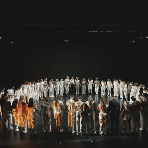 Duża grupa tancerzy stojąca w kole na deskach teatru, na czarnym tle.