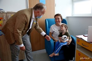 W pokoju szpitalnym prezydent Gdyni wita się z dzieckiem siedzącym na kolanach mamy, która siedzi w fotelu.