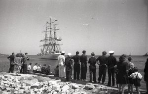 Negatyw czarno-biały z 1946 roku przedstawia tłum ludzi na zniszczonym nabrzeżu, którzy żegnają wypływający w rejs statek „Dar Pomorza”.  W głębi widoczna rufa płynącego żaglowca. Fotografia pochodzi z archiwum cyfrowego Muzeum Miasta Gdyni // fot. Edmund Zdanowski