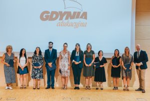 Na scenie w rzędzie grupa ludzi, w tle ekran rzutnika z logiem Gdyni edukacyjnej na białym tle 