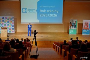 Widok auli z góry, na scenie kobieta w niebieskim żakiecie mówiąca do mikrofonu, który trzyma w rękach