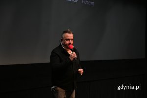 Mężczyzna w ciemnym ubraniu z mikrofonem na tle kinowego ekranu