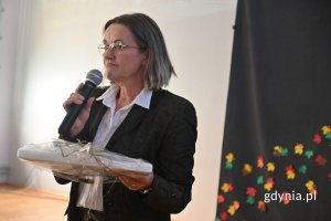 Stojąca kobieta mówiąca do mikrofonu w szkolnej hali sportowej
