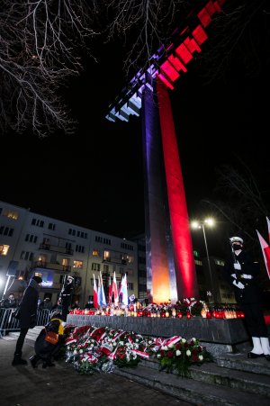Pomnik Ofiar Grudnia '70 na placu Wolnej Polski . Krzyż oświetlony w barwach narodowych. Przed pomnikiem dwie kobiety - jedna składa kwiaty, druga stoi. Po obu stronach pomnika posterunek honorowy złożony z marynarzy. Przed pomnikiem leży mnóstwo wieńców i wiązanek kwiatów. Płyta pomnika wypełniona jest zniczami // fot. Karol Stańczak