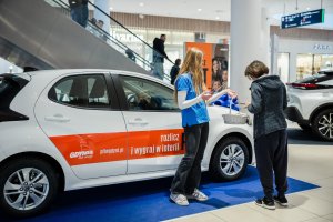 Kobieta w grantaowej kurtce przy aucie, hostesa podaje jej ulotkę z białego koszyka