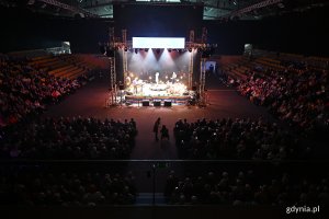 Hala Polsat Plus Arena Gdynia, scena, dekoracja ze stołem i krzesłami, aktorzy na scenie, publiczność dookoła, nad sceną wyświetlacz