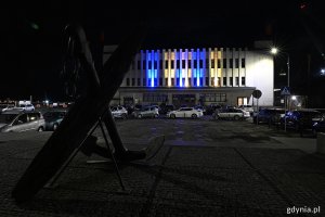 na zdjęciu budynek podświetlony w żółte i niebieskie pasy, na pierwszym planie kotwica