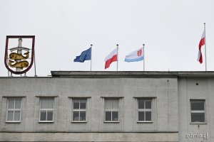 Flagi powiewające na budynku Urzędu Miasta Gdyni (od lewej): flaga Unii Europejskiej, flaga Polski, flaga Gdyni. Na maszcie biało-czerwona flaga. Po lewej stronie podświetlany herb Gdyni // fot. Michał Puszczewicz