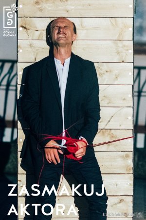 Grafika promująca wydarzenie Jubileusz 10-lecia Teatru Gdynia Główna. "Zasmakuj aktora". Mężczyzna w czarnym płaszczu i białej koszuli, z grymasem na twarzy. Ma związane ręce czerwonym sznurkiem. W prawym górnym rogu logo Teatru Gdynia Główna.