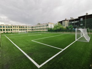 Na zdjęciu bosiko ze sztucznej trawy, nowe piłkochwyty z zielonej siatki, w tle budynek szkoły "tysiąclatki", a po prawej budynek starej drewnianej szkoły.