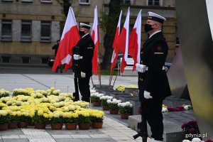 Posterunek honorowy przed pomnikiem Polski Morskiej z okazji Święta Niepodległości. Dwóch żołnierzy Marynarki Wojennej na tle biało-czerwonych flag. Na dole, po lewej stronie donice z żółtymi chryzantemami // fot. Michał Puszczewicz