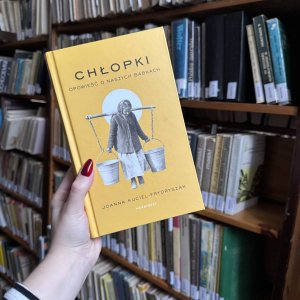 Kobieca dłoń trzymająca książkę „Chłopki. Opowieść o naszych babkach” na tle regału z książkami