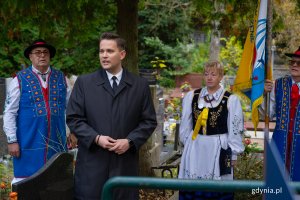 Mężczyzna w czarnym garniturze i para w regionalnym stroju nad grobem.