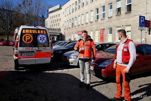 Dwaj mężczyxni w strojach medycznych stoją obok karetki pogotowia.