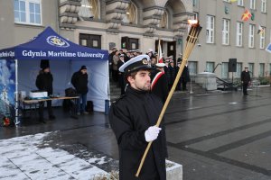 Student z pochodnią stoi na tle gmachu głównego Uniwersytetu Morskiego. Na schodach prowadzących do budynku stoi senat uczelni i goście. Za studentem, po lewej stronie stoi namiot z nazwą uczelni // fot. UMG