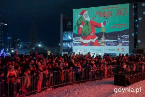 Gdynianie zgromadzeni na placu Grunwaldzkim podczas zabawy mikołajkowej. Za nimi, na ścianie budynku Teatru Muzycznego, wyświetlony plakat promujący wydarzenie 