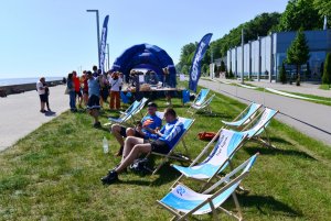 Bulwar, na środku trawa, na której stoją lerzaki w biało-błękitnych barwach, w tle niebieski dmuchany namiot Gdynia Sport. Na leżakach odpoczywa dwóch biegaczy, w tle stoisko ze słodkim kiermaszem III LO, przy nim grupka ludzi