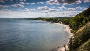 Panorama. Morze, częściowo zakryte chmurami niebo oraz plaża, molo i lasy. 