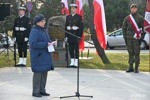 Na pierwszym planie, przy mikrofonie: Helga Łukaszewska, prezes gdyńskiego koła Światowego Związku Żołnierzy Armii Krajowej, podczas uroczystości z okazji 80. rocznicy utworzenia Armii Krajowej. Na drugim planie posterunek honorowy przy pomniku Armii Krajowej na skwerze Kościuszki. W tle widać biało-czerwone flagi  // fot. Magdalena Czernek