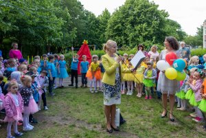 Grupy przedszkolne, w tle statyw na którym czeka na odsłonięcie pod czerwoną kotarą element metaloplastyki z nazwą, naokoło kolorowo ubrane dzieci