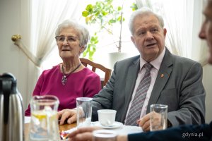 Starsza kobieta w różowej bluzce z siwymi włosami oraz starszy mężczyzna w szarym garniturze, siedzący przy stole 