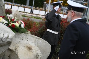 Mężczyźni w mundurach obok pomnika z szarych kamieni, na którym leżą białe i czerwone kwiaty