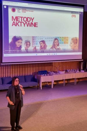 Kobieta przemawiajacy przed widownią z mikrofonem w ręku, wdalszym planie prezentacja