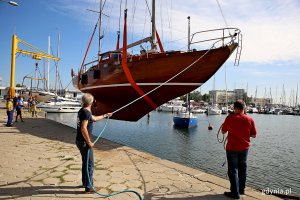 Mężczyźni asekurują linami jacht opuszczany dźwigiem do basenu w marinie.