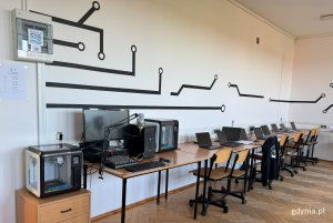 Sala ze stołami w rzędzie pod ścianą, na których stoją laptopy, w pierwszym planie drukarka 3d, dalej radiowęzeł, kolejna drukarka i laptopy