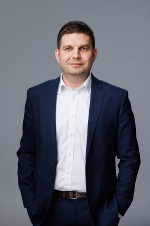 Zdjęcie portertowe: Maciej Zielonka, Wiceprezydent Miasta Gdyni, w garniturze i białej koszuli, na szarym tle. 
