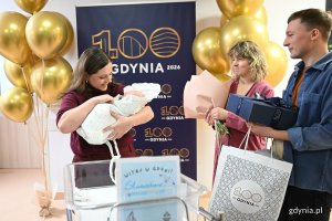 Kobieta trzyma na rękach noworodka, obok mężczyzna i kobieta z umpominkami w dłoniach