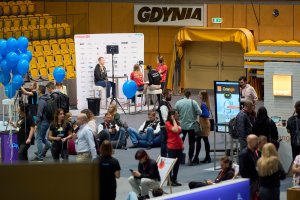 Grupa siedzących, stojących i rozmawiających osób w hali