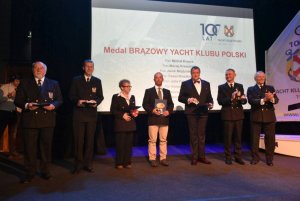 Grupa osób stojących na scenie i mających za plecami biały ekran