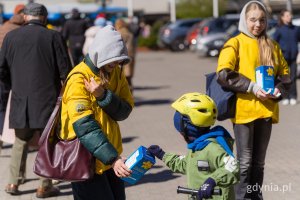 Mały chłopiec na rowerku wrzuca pieniądze do skarbonki, kt&oacute;rą trzyma wolonatariuszka w ż&oacute;łtej koszulce