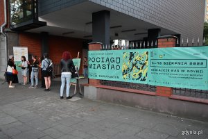 Wejście na patio Konsulatu Kultury, gdzie odbył się pokaz jednego z przedstawień inaugurujących festiwal „Pociąg do Miasta”. Po lewej - grupa osób stojących przed wejściem. Po prawej, na płocie - zielony baner promujący festiwal // fot. Michał Puszczewicz