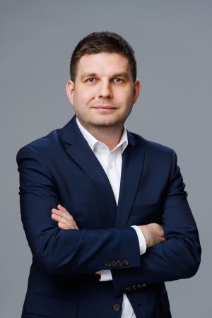 Zdjęcie portertowe: Maciej Zielonka, Wiceprezydent Miasta Gdyni, w garniturze i białej koszuli, na szarym tle. 