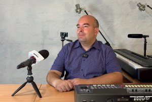 Mężczyzna siedzący przy stole, ustawiony po skośie przed mikrofonem w pomieszczeniu do nagrań. Gotowy do wypowiedzi.